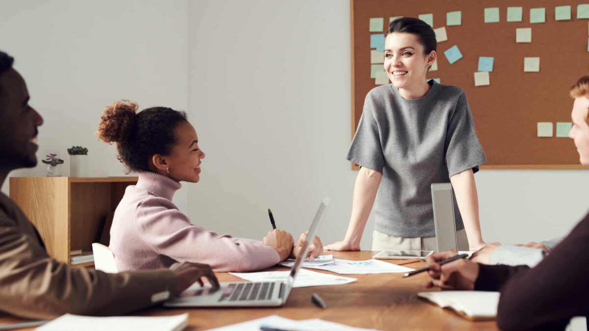 Diversiteit - Vrouw staat aan het hoofd van een tafel tijdens een vergadering en brainstorm waar nog 3 andere mensen bij aanwezig zijn